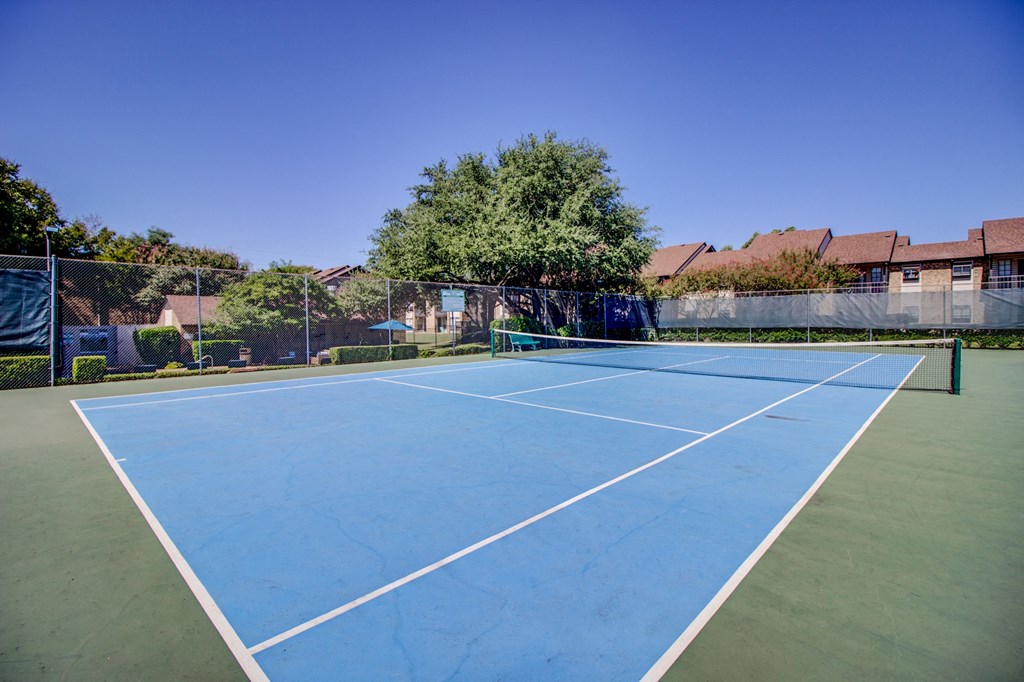 a blue and green tennis court with a fence and houses in the background