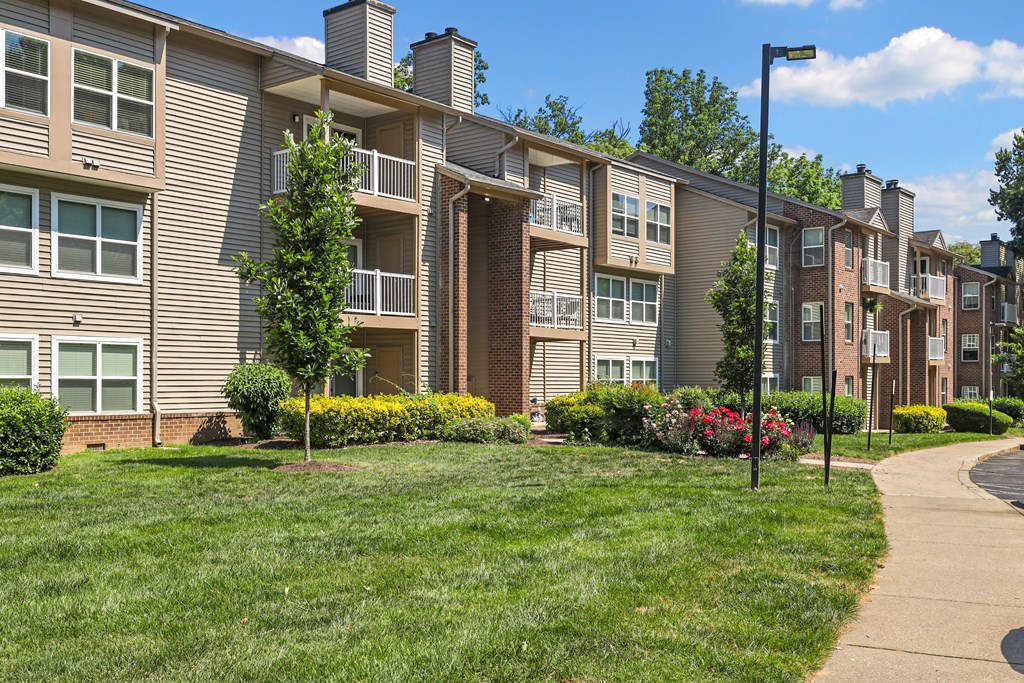 a grassy area with trees and bushes in front of an apartment building
