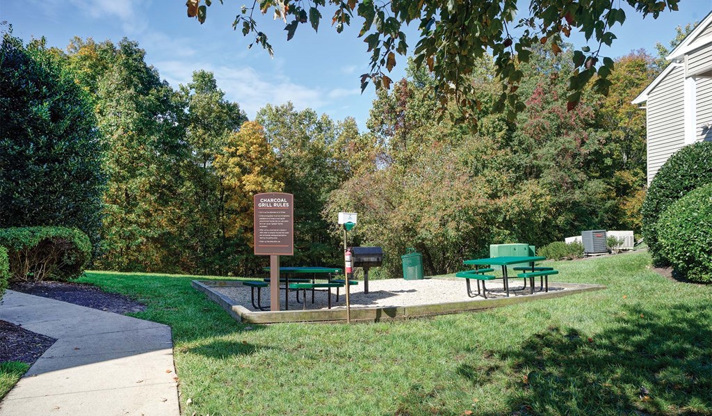 Outdoor grilling area with two green picnic tables surrounded by grass and trees. 