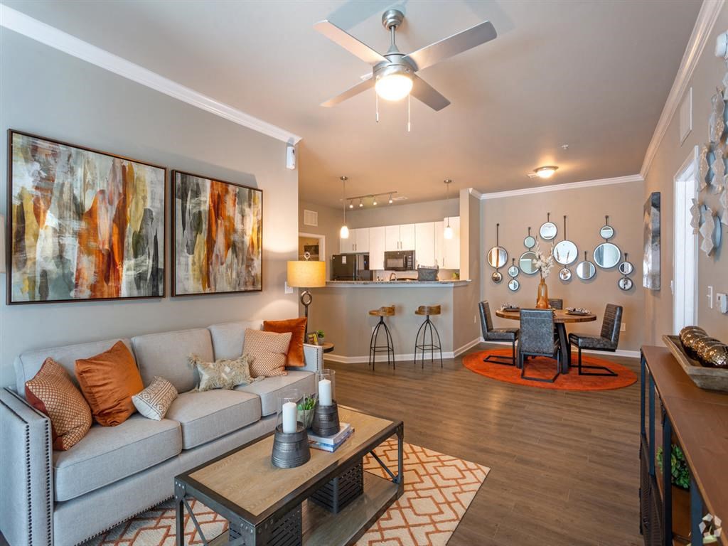 Spacious living room with ample seating, ceiling fan, wood-grain flooring, and view of kitchen at Lullwater at Blair Stone apartments for rent