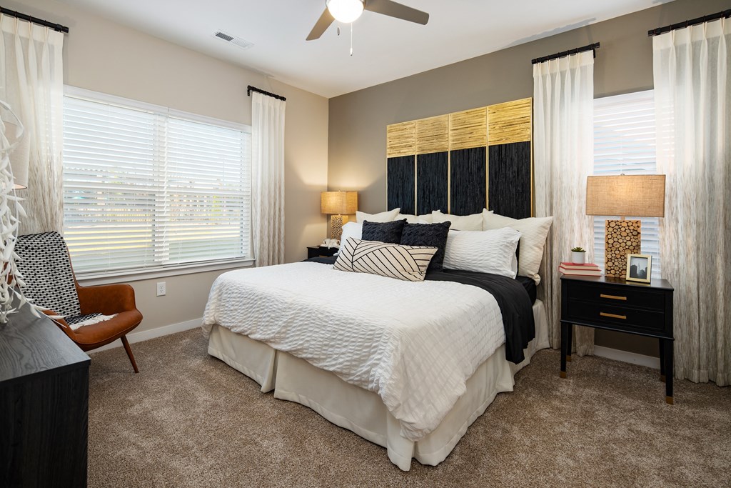 Furnished bedroom with queen size bed, two nightstands, carpet flooring, windows, and high ceiling at The Alexandria apartments in Madison, AL Furnished bedroom with queen size bed, two nightstands, carpet flooring, windows, and high ceiling at The Alexandria apartments in Madison, AL