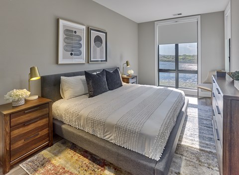A bedroom with a large bed, a nightstand with a lamp, and a view of the beach through the window.
