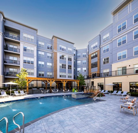 A large swimming pool surrounded by a wooden deck and lounge chairs in front of apartment buildings.