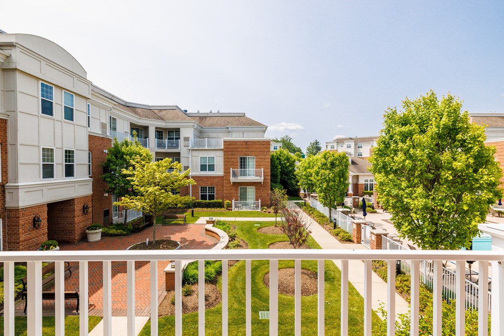 A white balcony overlooks a courtyard with a brick building and a tree.