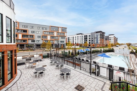 A patio with tables and chairs overlooks a pool and buildings.