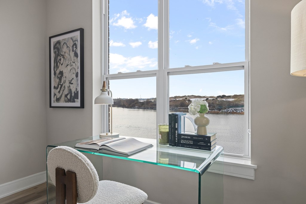 A room with a chair, a table with books and a lamp, and a window with a view of the water.