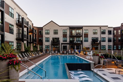 A swimming pool is surrounded by lounge chairs and umbrellas in front of apartment buildings.