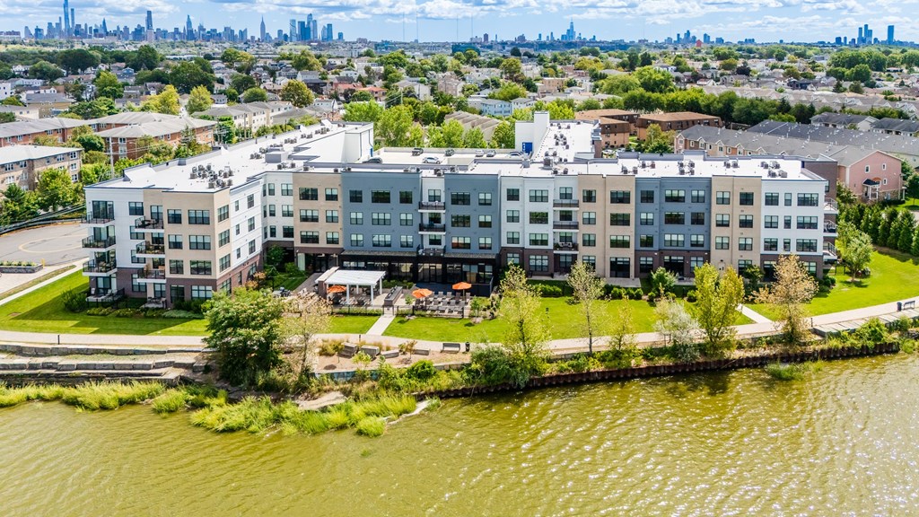 A large apartment complex sits on the edge of a body of water with a city skyline in the background.