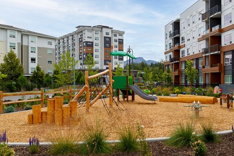 A playground with a slide, swings, and climbing structures in the foreground with apartment buildings in the background.
