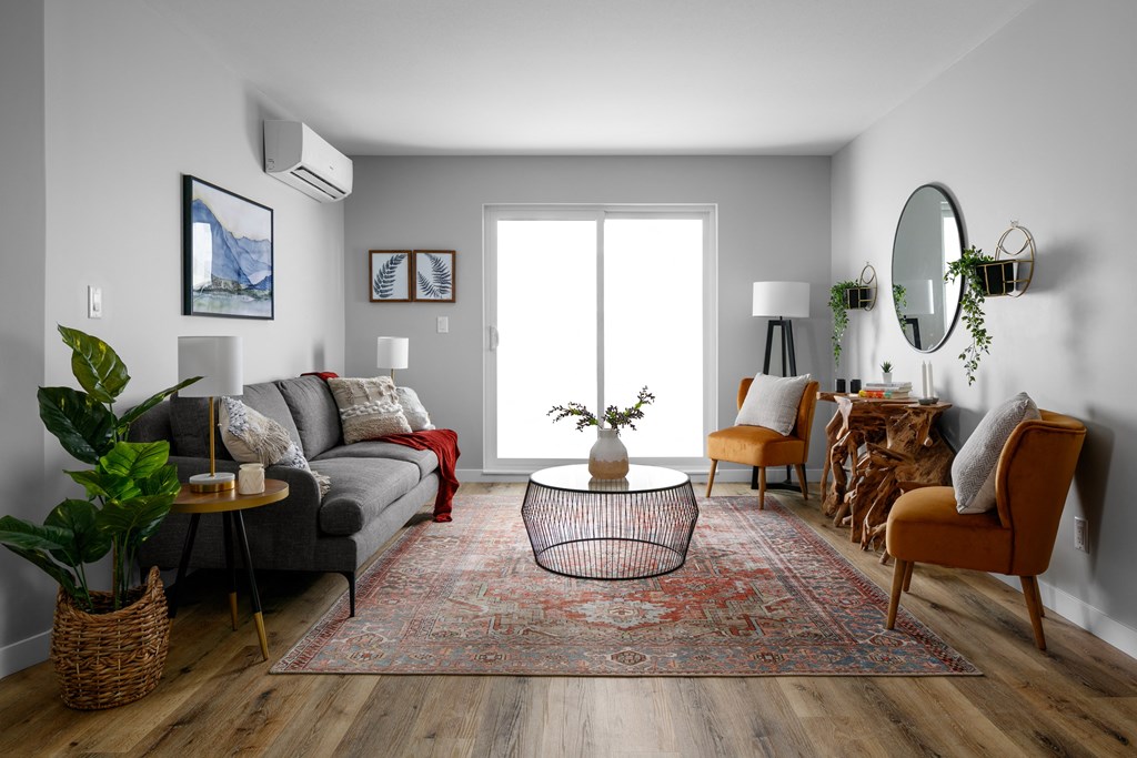A living room with a grey couch, a red throw, a round table with a plant on it, a chair, a rug, and a mirror on the wall.