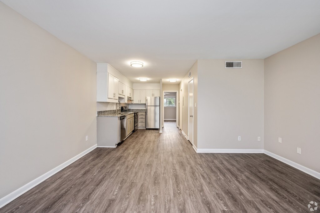 A spacious room with wood flooring and a kitchen area in the background.