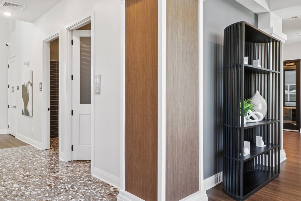 A black shelf with a plant on top is in the foreground of a hallway with white walls and a patterned floor.