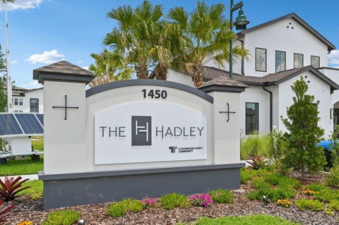 The Hadley sign is in front of a white building with a palm tree. at The Hadley - North Port, FL Apartments, North Port, FL