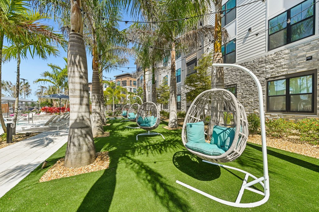 A white swing chair with a green cushion is in the foreground of a sunny outdoor area with palm trees and a building in the background. at The Concord Luxury Apartments, Florida
