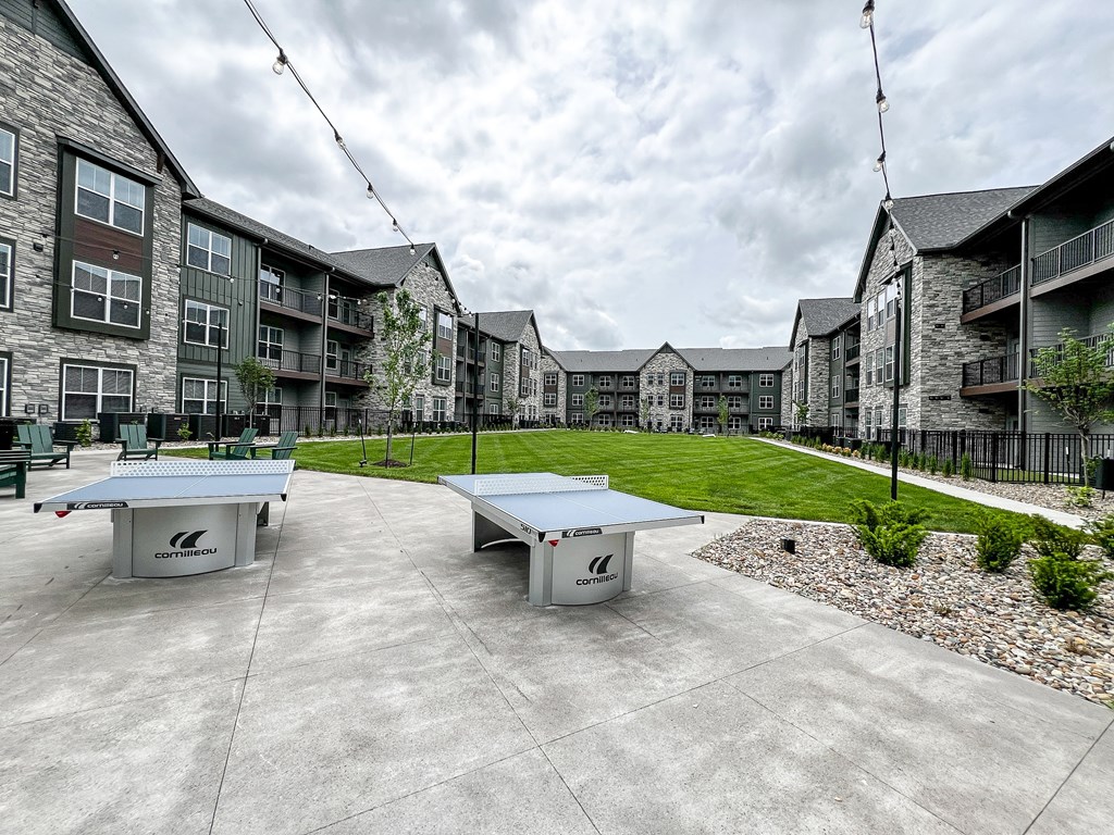 an outdoor patio with two ping pong tables in front of an apartment building at The Depot, Raymore, MO, 64083