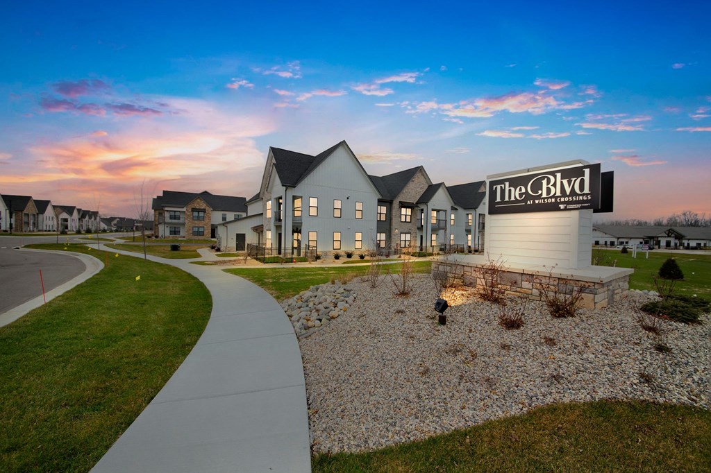 an exterior view of the residences at dusk with a sidewalk and grass at The BLVD at Wilson Crossings, Wyoming, MI 49418