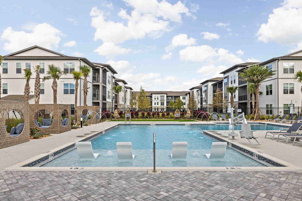A swimming pool surrounded by lounge chairs and palm trees in front of apartment buildings.