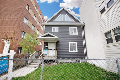 a gray house with a gray roof behind a chain link fence and next to a brick building