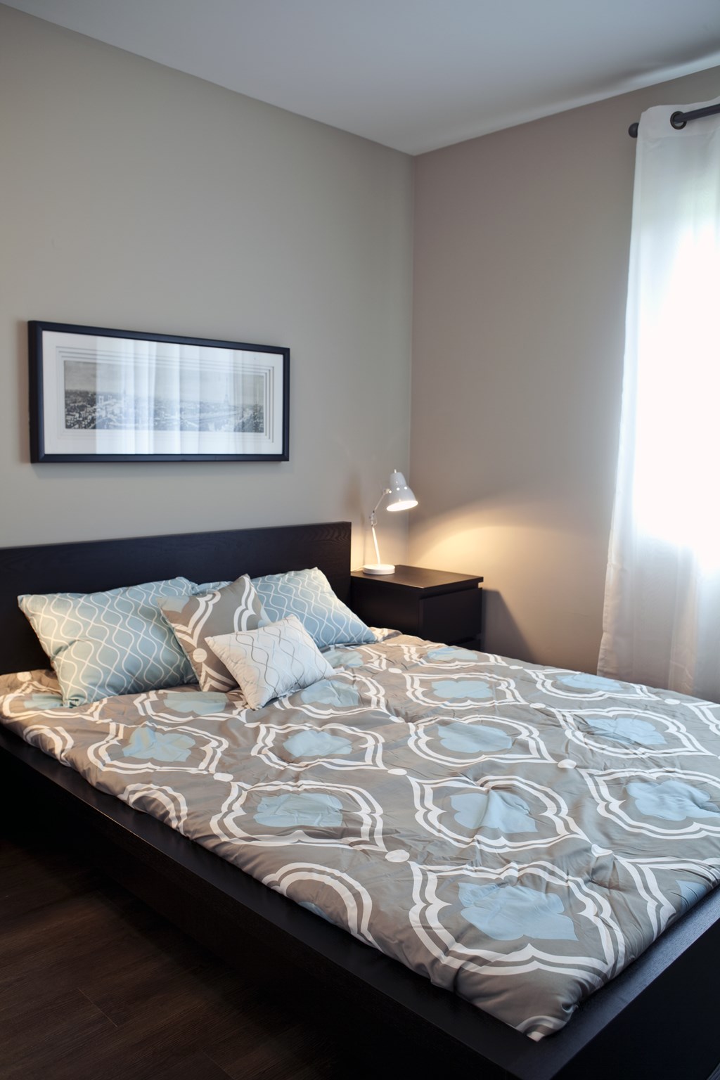 A bed with a grey and white patterned comforter and two blue patterned pillows sits in a room with a framed picture on the wall and a lamp on a side table.