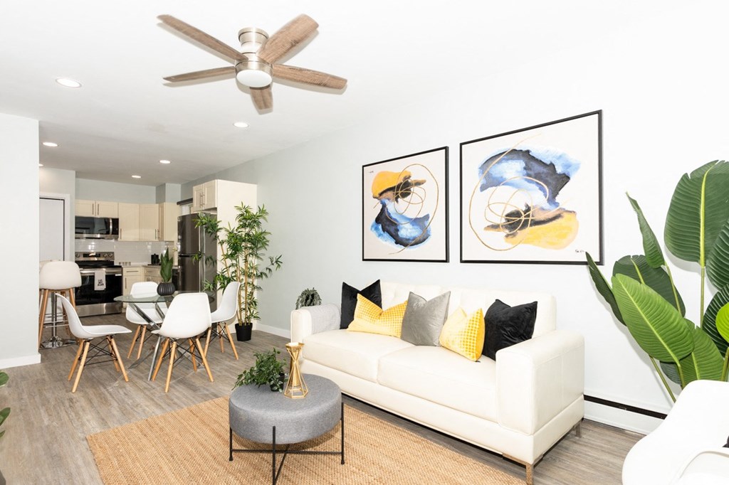 A living room with a white couch, a grey ottoman, and a ceiling fan.