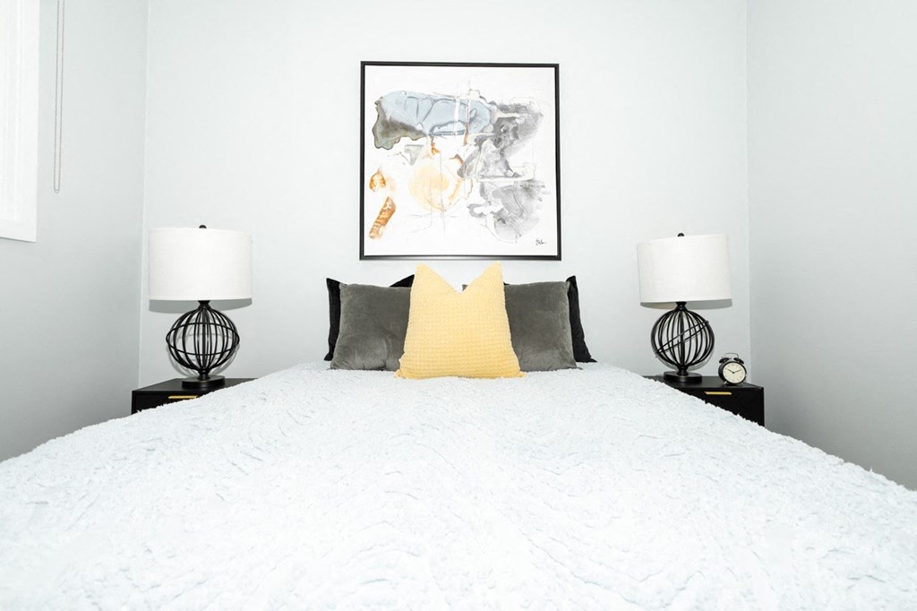 A bed with a white comforter and a yellow pillow is flanked by two black lamps.
