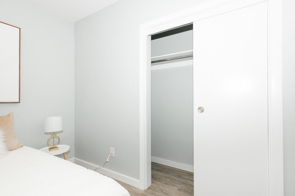 A white bedroom with a bed, nightstand, and a closet with a glass door.