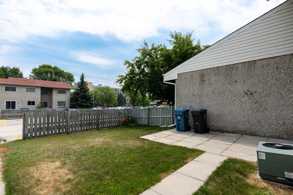 A backyard with a fence, a trash can, and a building in the background.