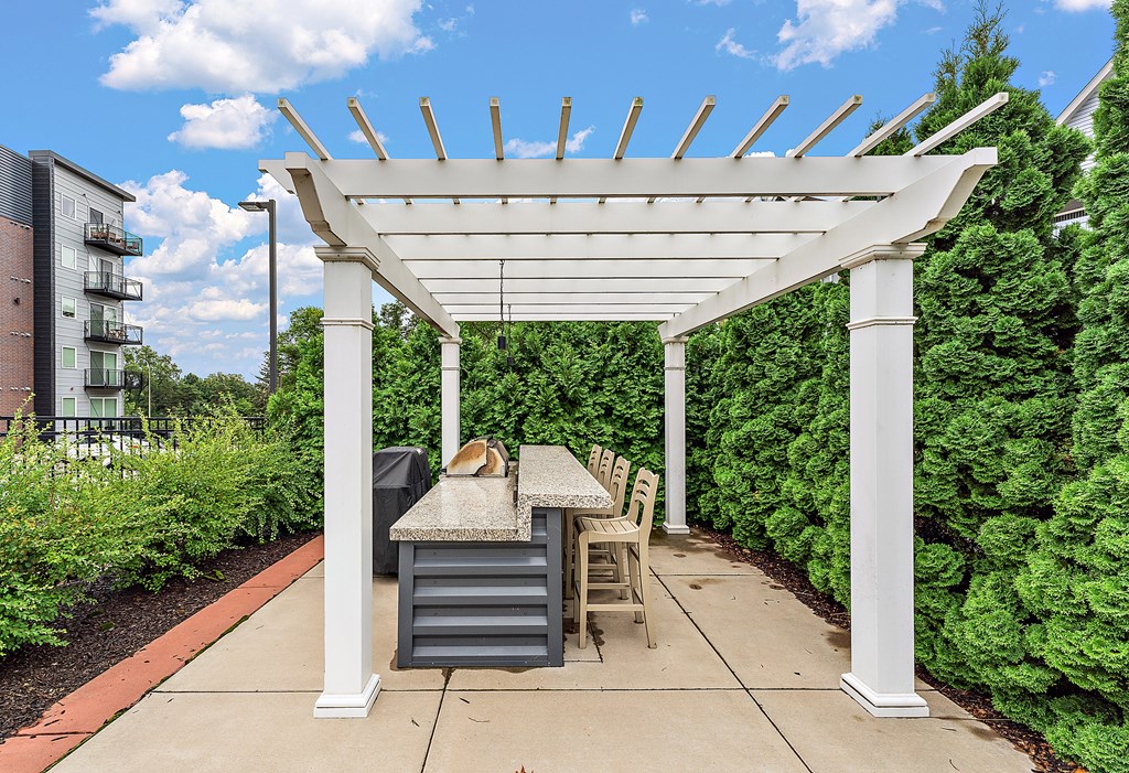 A white pergola with a table and chairs is surrounded by green shrubbery.