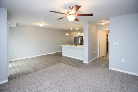 A spacious living room with a ceiling fan and a kitchenette in the background.