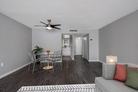 A living room with a grey couch, a dining table with chairs, a ceiling fan, and a rug on the floor.