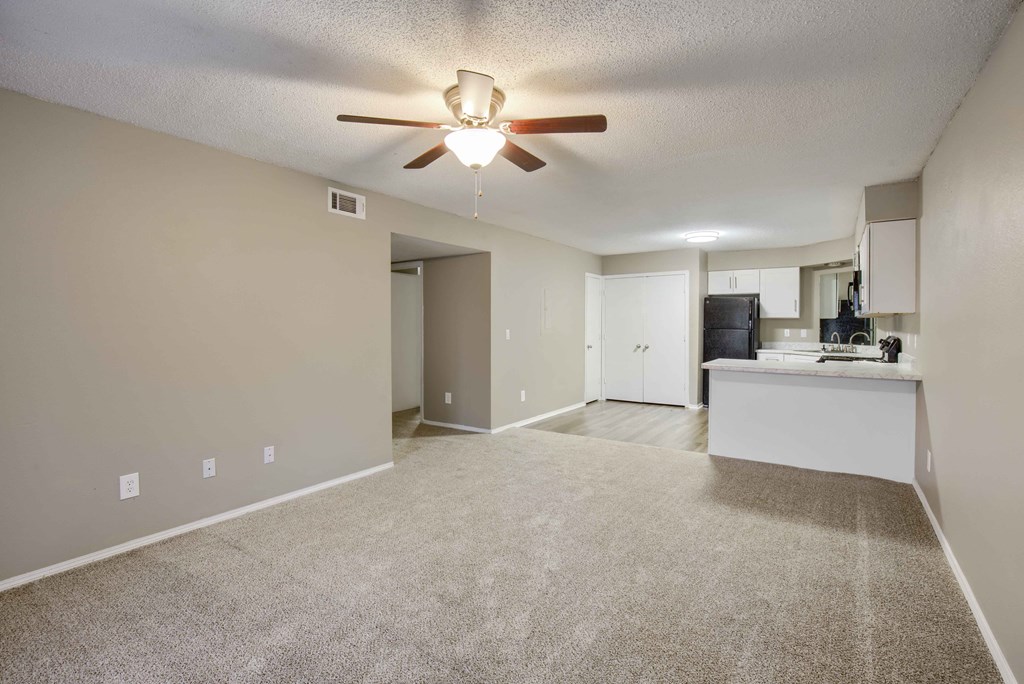 an empty living room with a ceiling fan and a kitchen in the background