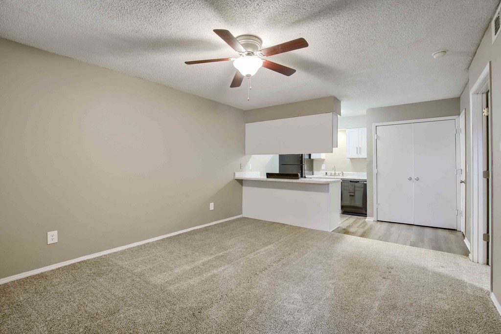 an empty room with a ceiling fan and a kitchen in the background