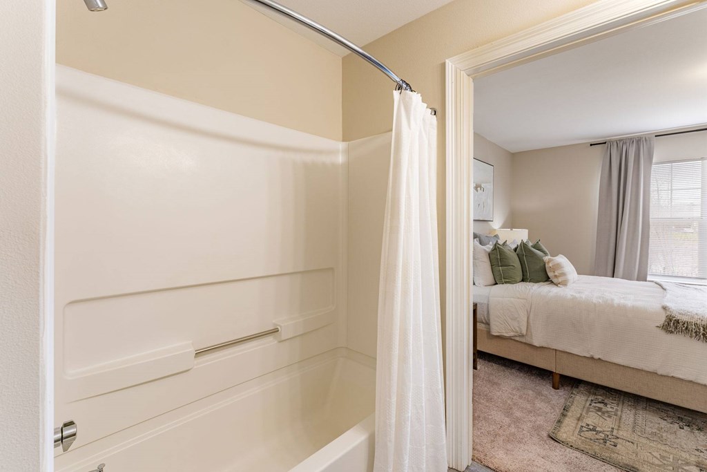 A white bathroom with a shower and a bedroom in the background.