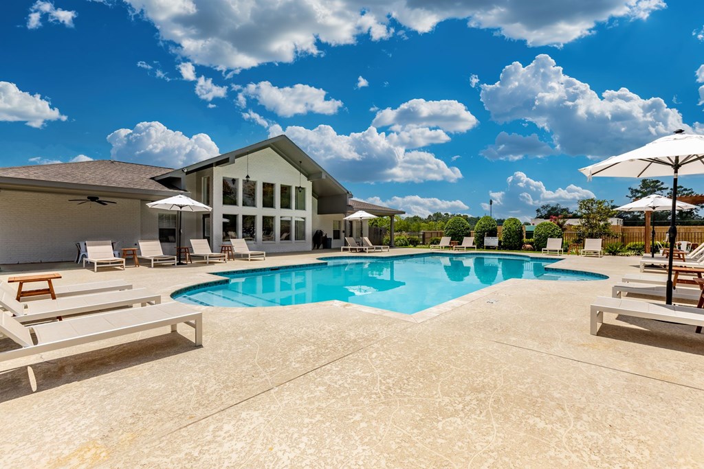 A large swimming pool in front of a house with a patio and lounge chairs.