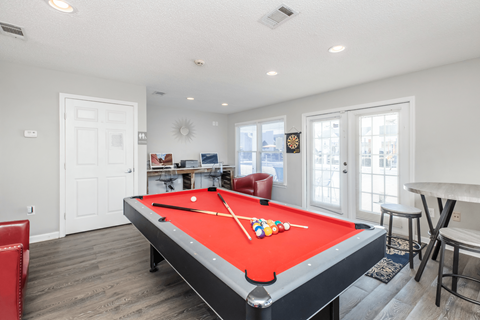 A pool table with a red cloth and white cues is in the center of a room with a white door and a window.