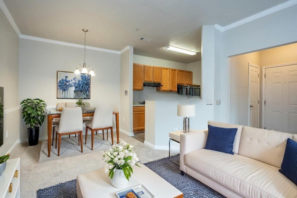 A living room with a white couch, a dining table with chairs, and a kitchen area in the background.
