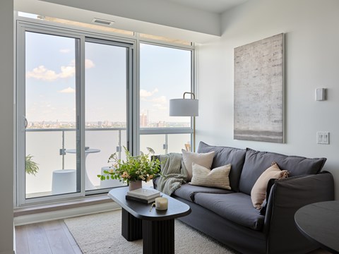 A living room with a grey couch and a large window overlooking the city.
