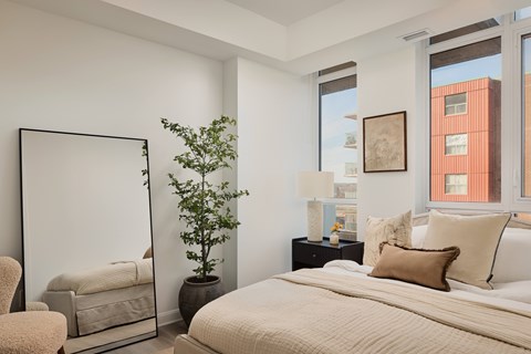 A bedroom with a large bed, a chair, a potted plant, and a mirror.