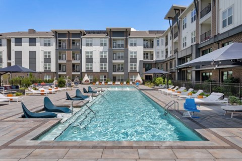 A large swimming pool surrounded by lounge chairs and umbrellas in front of apartment buildings.