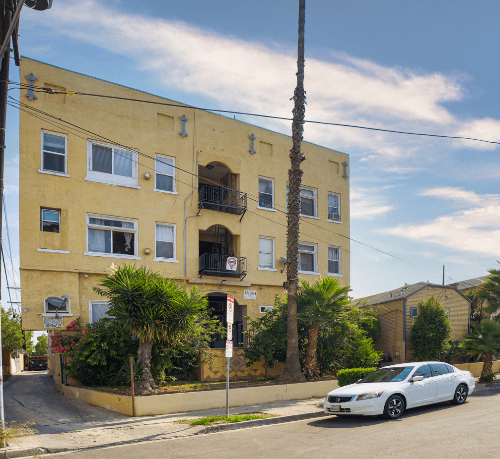 A white car is parked in front of a yellow building with a balcony.