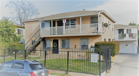 A car is parked in front of a two-story apartment building with a black fence.