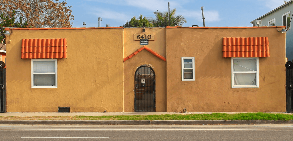 A tan building with a black door and two windows with red awnings.