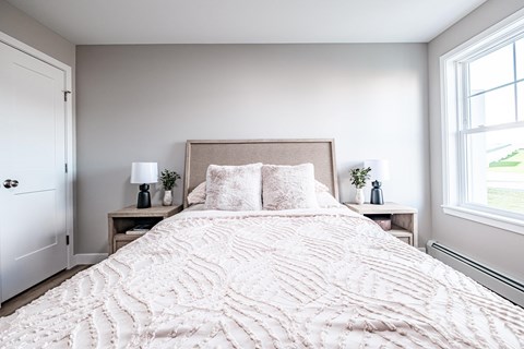 A bed with a white comforter and pillows is in the center of a bedroom with a white door and window.