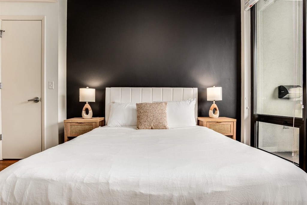A bed with white bedding and two pillows sits in a room with a white door and a black wall.