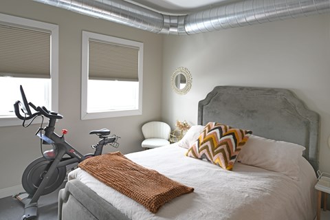 A bedroom with a bed, a grey headboard, a bicycle, and a window with blinds.