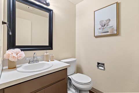 A bathroom with a toilet, sink, mirror, and a picture on the wall.