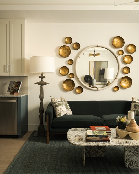A living room with a black couch, a round mirror with gold circles, and a table with a lamp and a plant.