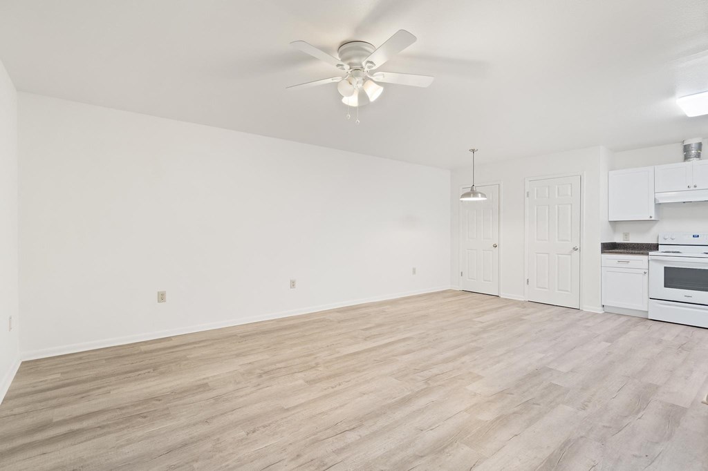 an empty living room with a ceiling fan and a kitchen in the background