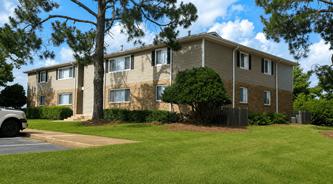 A building with a brown and beige exterior is surrounded by a green lawn and trees.