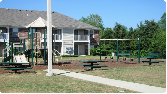 A playground with a slide, swings, and picnic tables in front of apartment buildings.